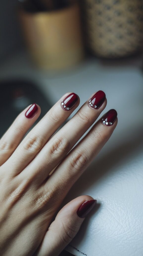 A hand with neatly manicured short nails is shown. The nails are painted a deep red color, with small silver beaded accents near the cuticles on a couple of the nails. The background includes a white surface and blurred objects, offering stylish ideas for an elegant look.