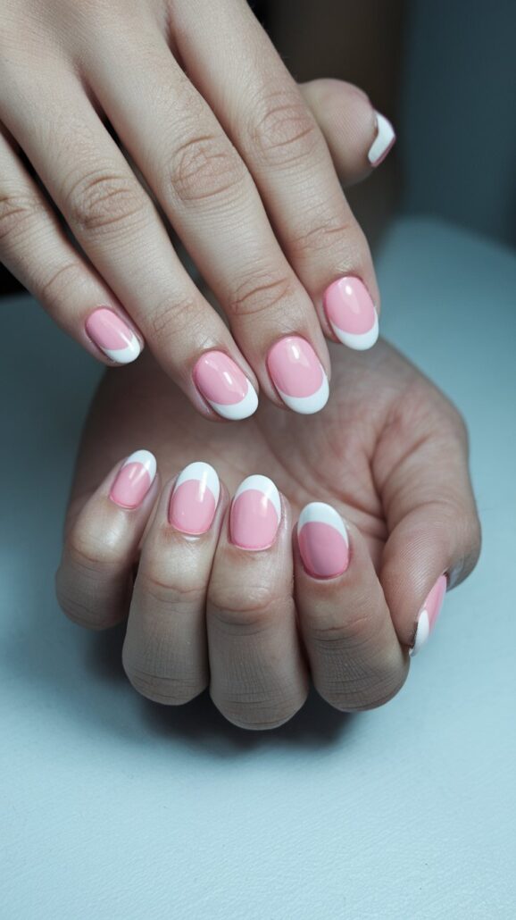 A close-up of hands flaunting a French manicure on short nails, with a pink base and white tips. The nails are neatly polished, exuding stylish ideas against a gray backdrop. The skin appears smooth and well-maintained.
