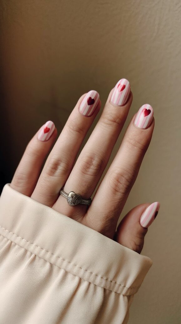 A hand with manicured nails showcases a stylish idea featuring a design of red hearts and vertical pink stripes, perfect for cruise nails. A ring with a heart motif adorns the middle finger, while the sleeve of a beige garment peeks through.