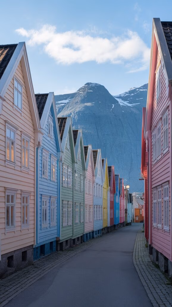 A row of colorful wooden houses lines the narrow street, their pastel shades of pink, blue, green, and yellow adding charm to the scene. A rocky mountain provides a stunning backdrop under a partly cloudy sky, making it a perfect destination to explore on your Northern Europe cruise.