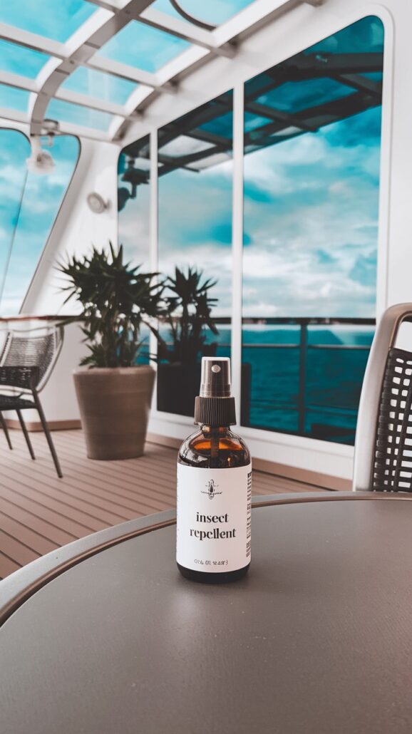A bottle of essential insect repellent sits on a round table on a ship's deck, surrounded by wicker chairs and a potted plant. Through the glass panels, the ocean stretches out beneath a partly cloudy sky—a perfect scene for a Bahamas cruise informed by smart packing tips.