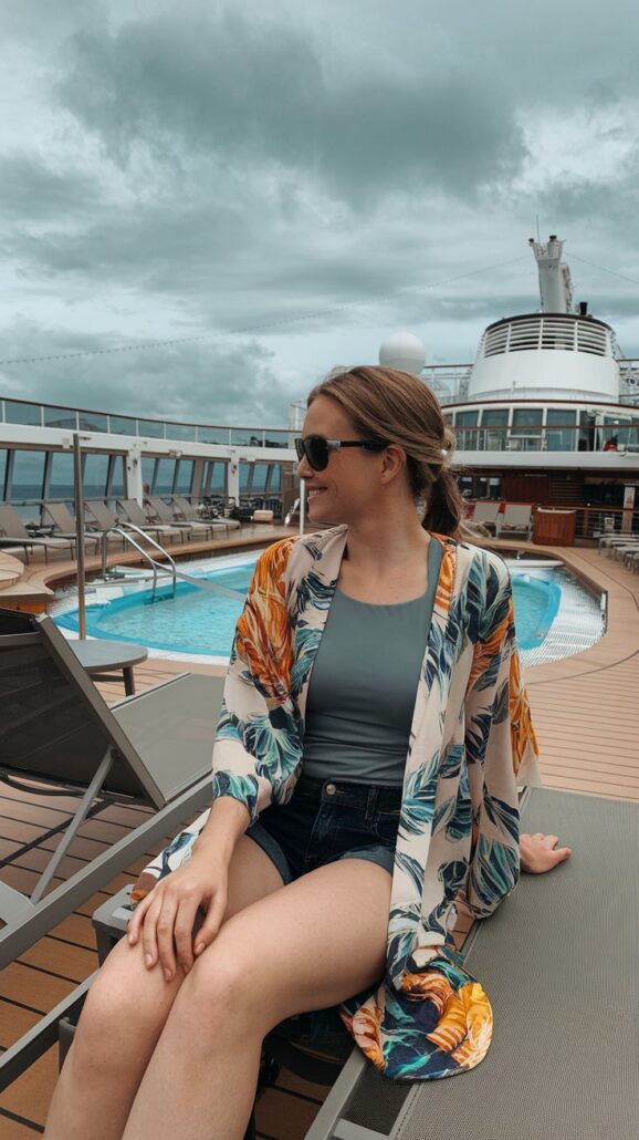 A person exudes chill style, donning sunglasses, a floral cover-up, a green top, and denim shorts as they relax on a deck chair by a pool on the cruise ship. The cloudy sky and array of deck chairs in the background complete this effortless women fashion scene.