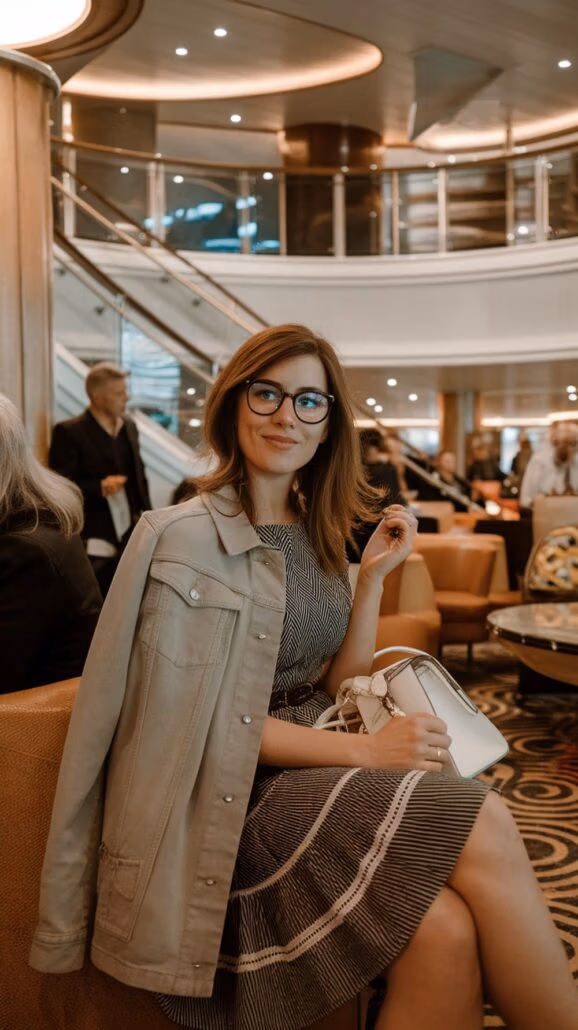 A woman with glasses is seated in a stylish indoor setting, embodying cozy outfit ideas. She wears a striped dress with a light-colored jacket draped over her shoulders, holding a small handbag and smiling warmly at the camera.