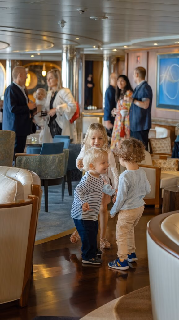 Three children stand together on a polished floor in a room with chairs and soft lighting, reminiscent of the cozy ambiance found on family-friendly Caribbean cruises. Moms and other adults chat in the background, some holding drinks in what seems to be a lounge or reception area.