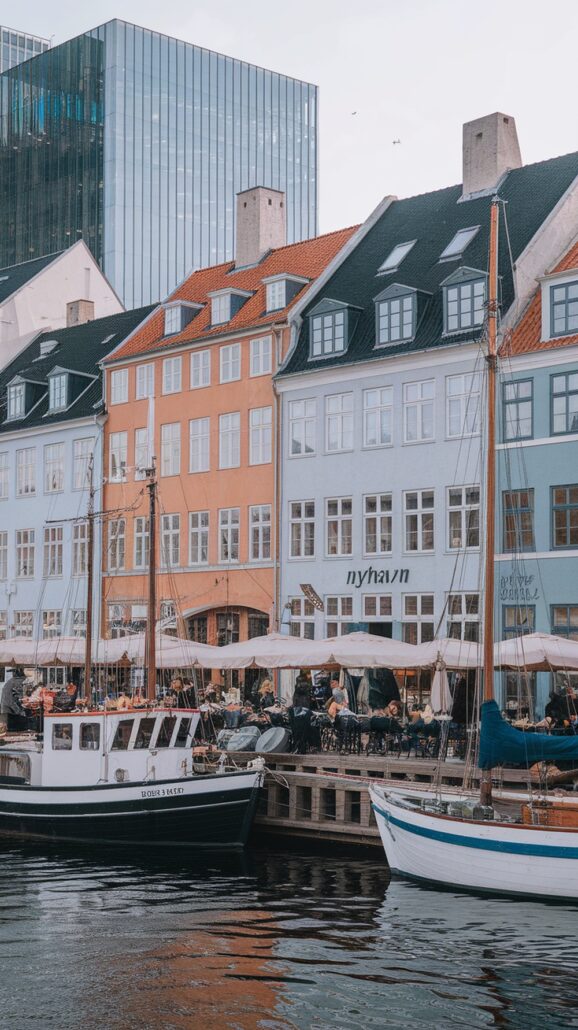 Explore the incredible destination of Nyhavn in Copenhagen, where colorful buildings line the canal. Boats bob gently in the water near a bustling outdoor café. The overcast sky casts a unique charm, with modern glass structures providing a stunning contrast in this Northern European gem.