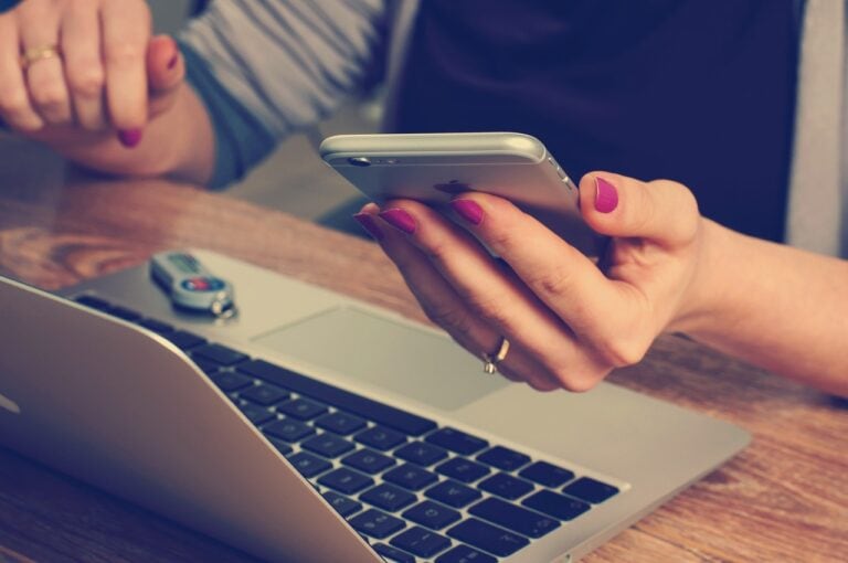 A person with pink nail polish holds a smartphone in one hand and rests the other on a laptop keyboard, possibly plotting their next move like a savvy scam artist. A USB flash drive is inserted into the laptop, all set against a wooden desk.