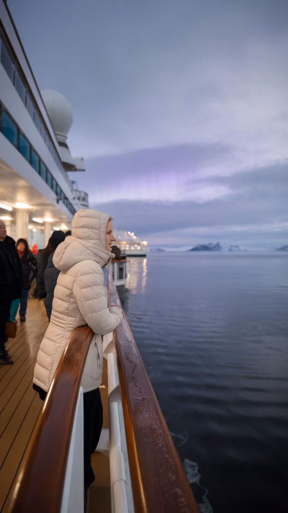 A person in a white jacket leans on the railing of a ship's deck, embracing the serene cruise adventure. Other individuals stand nearby as the sky, overcast with a hint of purple, stretches above distant mountains.