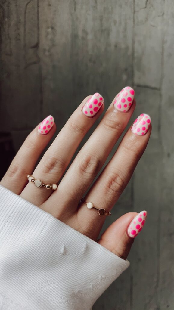 A hand flaunts pink and white polka dot nail art on short nails, paired with gold rings featuring small pearls. The person wears a white long-sleeved garment against a plain concrete wall, offering stylish ideas for cruise nails.