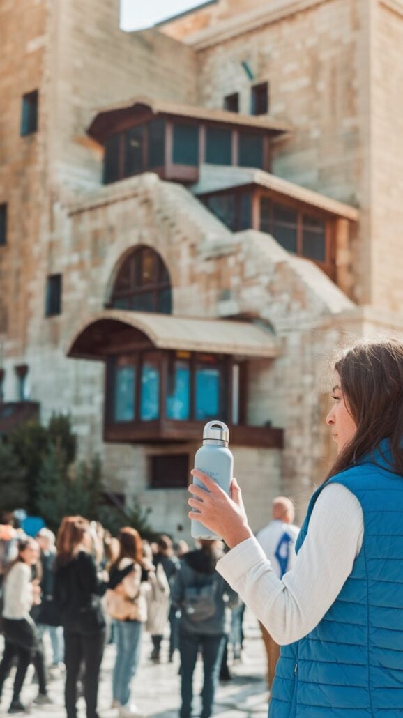 A woman in a blue vest, holding an essential item—a water bottle—stands in front of a large stone building with arched windows. A crowd gathers, some snapping photos under the sunny sky, adding vibrant energy to what feels like a stop on a Greek cruise.