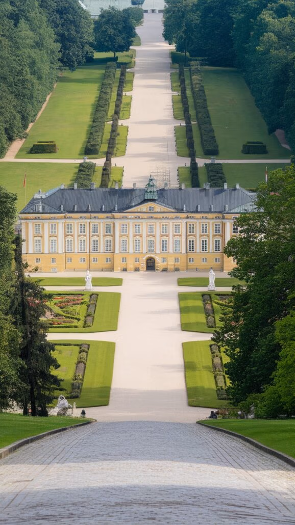 The image depicts a long avenue lined with lush greenery leading to a grand, yellow baroque-style palace. With its symmetrical facade and numerous windows, the palace invites you to explore its majesty. Manicured gardens and statues enhance this incredible destination.