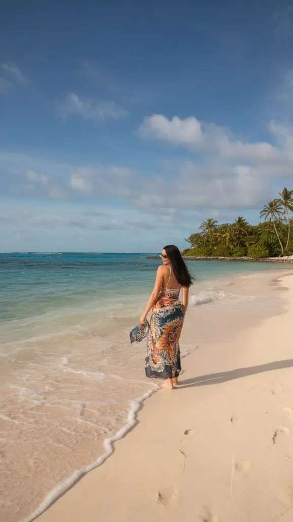 A woman with long dark hair walks barefoot along a sandy beach, wearing a colorful dress. She strolls near the shoreline where gentle waves lap at her feet, reminiscent of a relaxing Caribbean cruise. The ocean is to her left, framed by palm trees and lush greenery in the background.