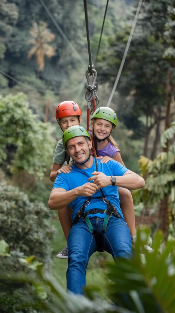 A person ziplining with two children in a lush, forested area is the essence of unforgettable family adventures. The adult, in a blue shirt and jeans, and the kids, with red and green helmets, are all harnessed together and smiling amidst the trees.