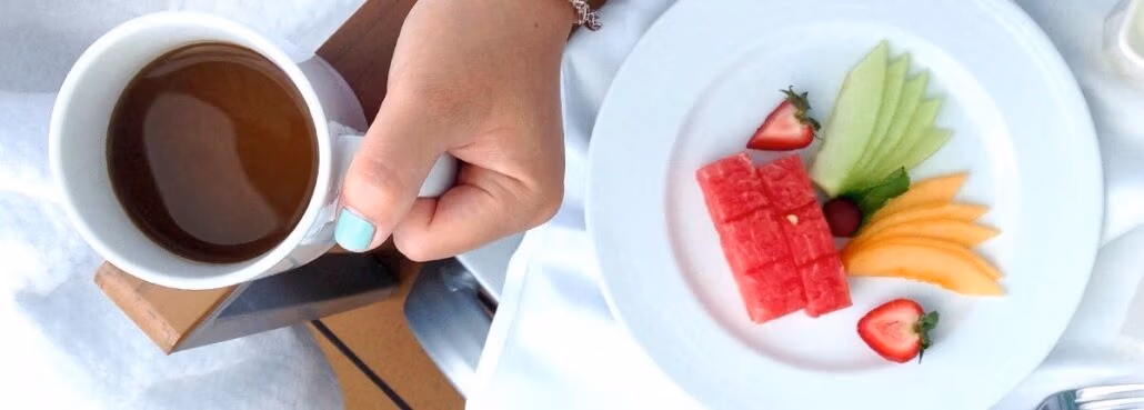 A person holds a cup of coffee in their left hand. On a white plate next to the cup, reminiscent of carnival cruise menus, are sliced watermelon, cantaloupe, honeydew, strawberries, and a grape, arranged neatly on a white tablecloth.