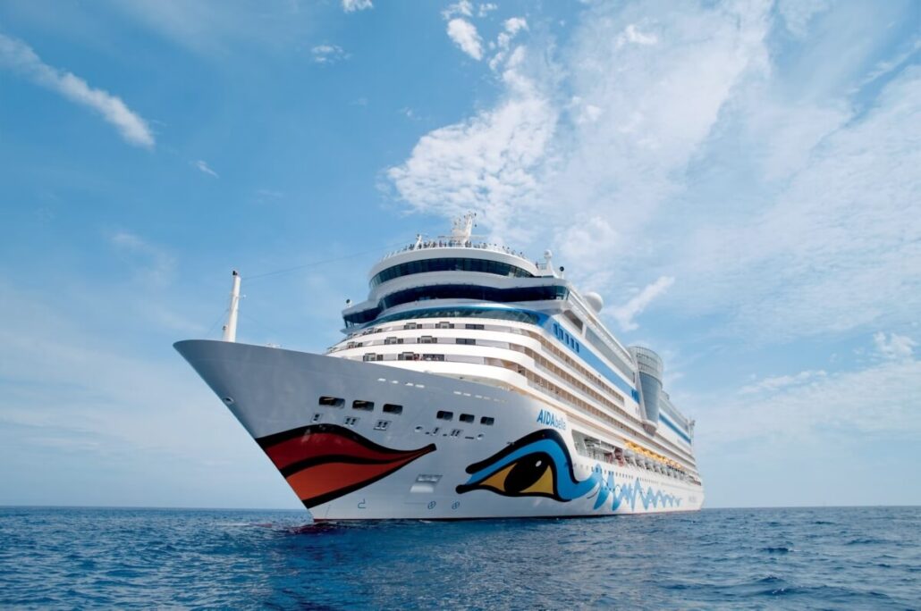 A large white cruise ship adorned with vibrant designs on the hull gracefully navigates the calm ocean under a partly cloudy blue sky. The bow, showcasing eye and wave graphics, bears the name "AIDA," embodying the spirit of an unforgettable cruise adventure.