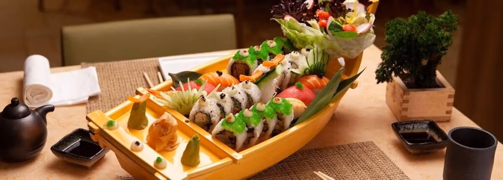 A wooden boat-shaped platter filled with assorted sushi, reminiscent of the exquisite offerings on carnival cruise menus, sits on a dining table. Nearby are a teapot, two dipping sauce dishes, napkins, and a potted plant.