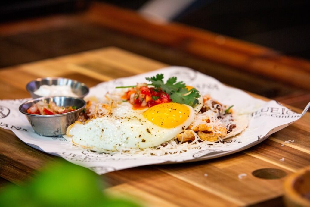 Amid whispers of an egg shortage, this dish on a wooden board showcases a fried egg sprinkled with pepper and shredded cheese, all beautifully garnished. It comes with salsa and a small metal cup of sauce, served elegantly on branded parchment paper—a treat worthy of the Carnival Cruise Line.
