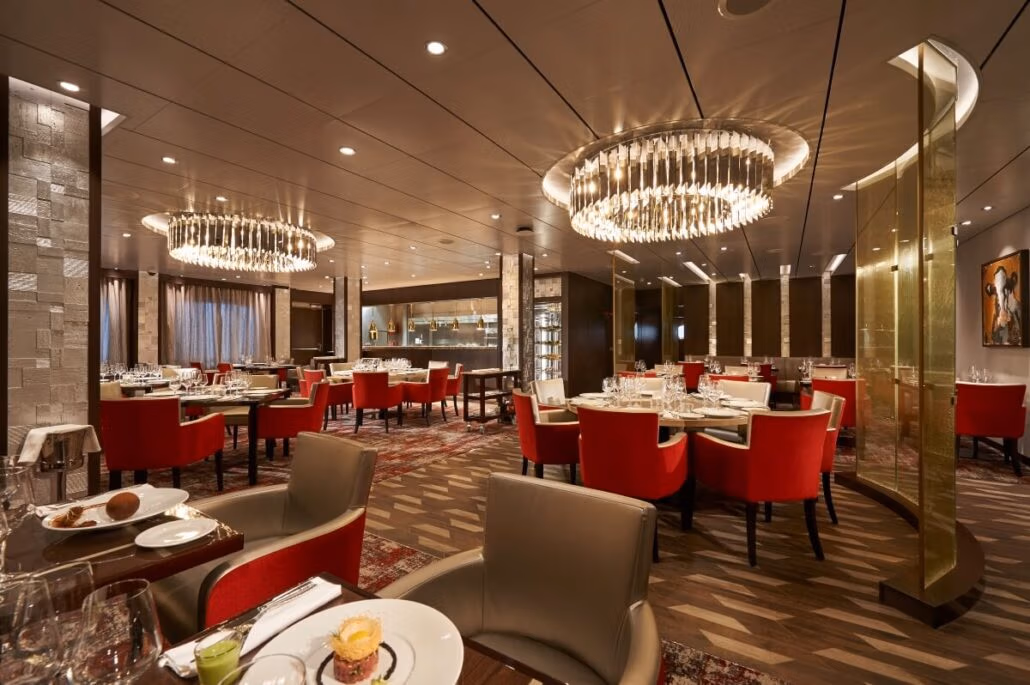 A luxurious restaurant interior with red upholstered chairs and white tablecloths evokes the elegance of an upscale dining experience found in carnival cruise menus. Two large, circular chandeliers hang from the ceiling, illuminating modern art-lined walls and a patterned carpeted floor.