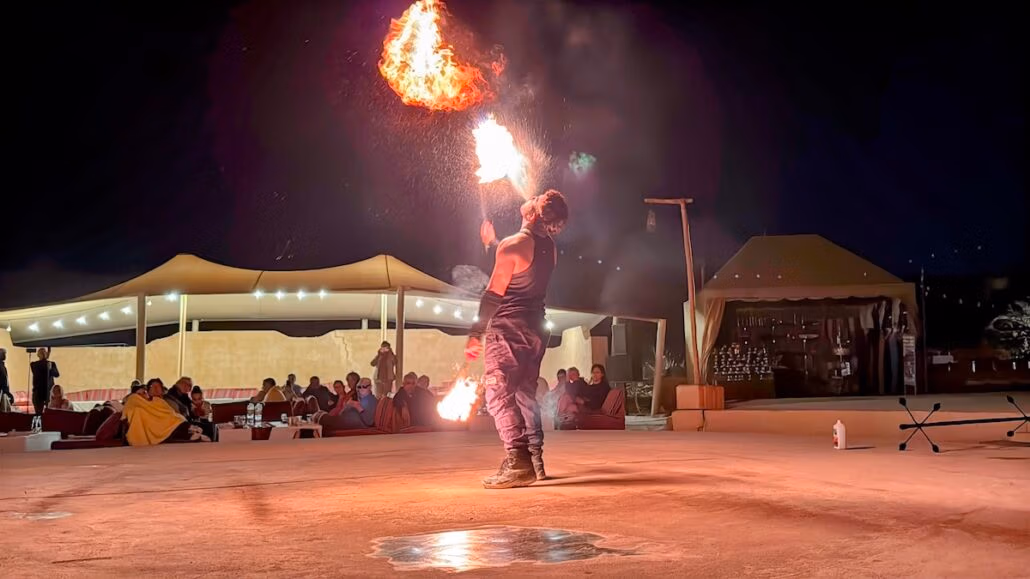 Under a starlit sky, as if on a Celestyal journey, a performer breathes fire into the night, captivating an audience seated under tents. The sandy open area serves as the perfect backdrop for this exhilarating spectacle, reminiscent of the magic found on a midsize ship adventure.
