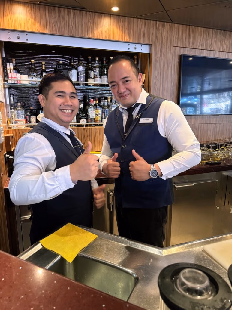 Two bartenders stand behind a bar counter, smiling and giving thumbs up. They're in sharp white shirts and blue vests, embodying the relaxed vibe of our Celestyal Journey trip. Behind them, shelves are stocked with bottles, while a TV on the wall to the right adds to this midsize ship's perfect charm.
