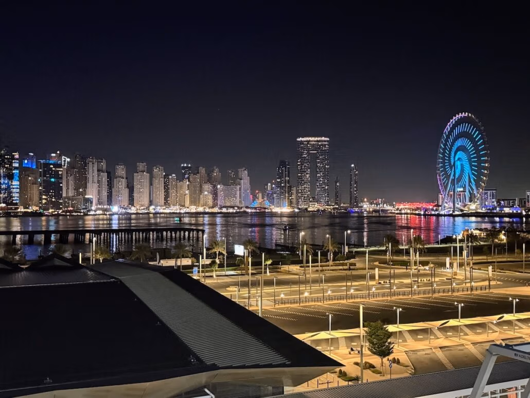 A night view of a cityscape boasts illuminated skyscrapers and a vibrant Ferris wheel reflecting on the water. In the foreground, reminiscent of a Celestyal Journey trip report, streetlights frame a sleek modern building that feels perfectly in tune with its surroundings.