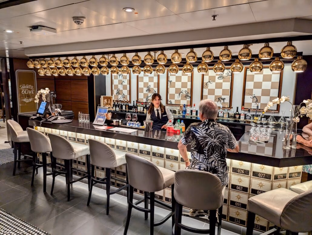 A person stands at a bar with a black counter, attended by a bartender. The ambiance mirrors the elegance of a Celestyal Journey, with golden hanging lights and chess-themed artwork on the decorative back wall, as bottles and glasses are artfully arranged across the surface.