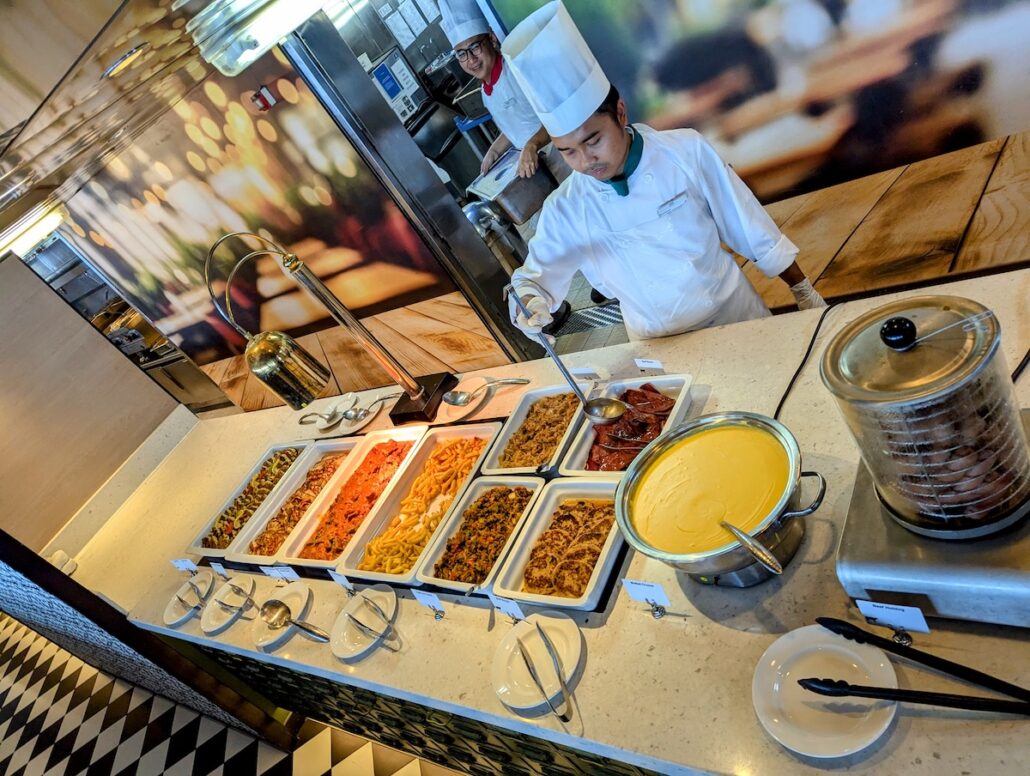 A chef in a white uniform and toque ladles food at a buffet counter on the Celestyal Journey mid-size ship. The counter features assorted dishes, bowls, and a large pot of yellow soup, embodying why this trip feels perfect amid the blurred restaurant decor.