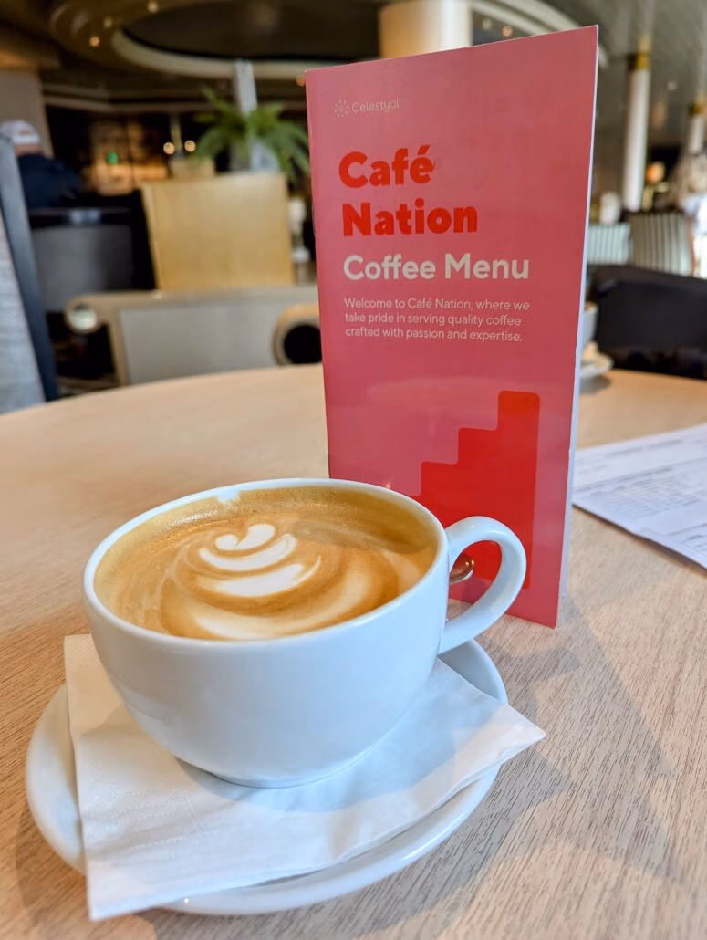 A cappuccino with latte art in a white cup on a saucer sits on a wooden table, much like the cozy setting you'd find during a Celestyal Journey. Behind it, a red menu card reads "Café Nation Coffee Menu." The blurred interior adds to the ship's perfect ambiance.