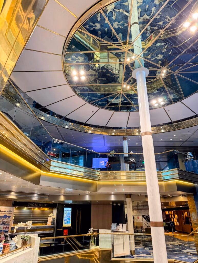 The image captures the stunning atrium of a Celestyal Journey ship, where a decorative circular ceiling depicts a starry sky. Multiple levels of glass balconies, bright overhead lighting, and an information desk on the lower floor highlight why this midsize ship feels perfect.