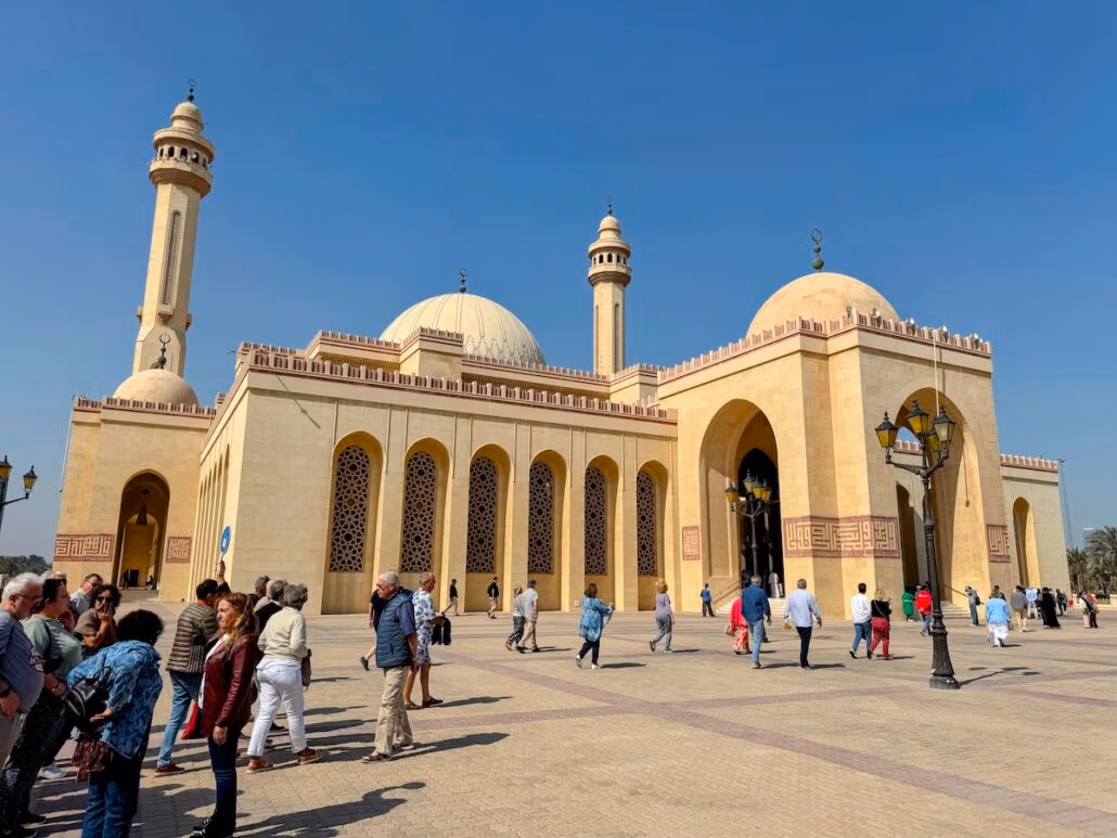People walk in front of a large mosque with beige stone walls, multiple domes, and tall minarets under a clear blue sky. Much like the charm found on the Celestyal Journey, this architectural wonder features ornate windows and arched entrances. Street lamps line the spacious courtyard.