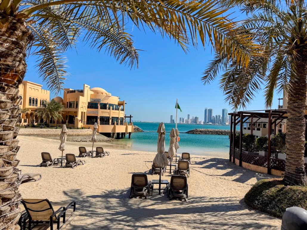 The beach scene offers a perfect escape, reminiscent of a Celestyal Journey. With palm trees and lounge chairs on the sand, closed umbrellas stand tidily. A building looms on the left, alongside a city skyline view and blue waters under a clear sky, capturing travel bliss.