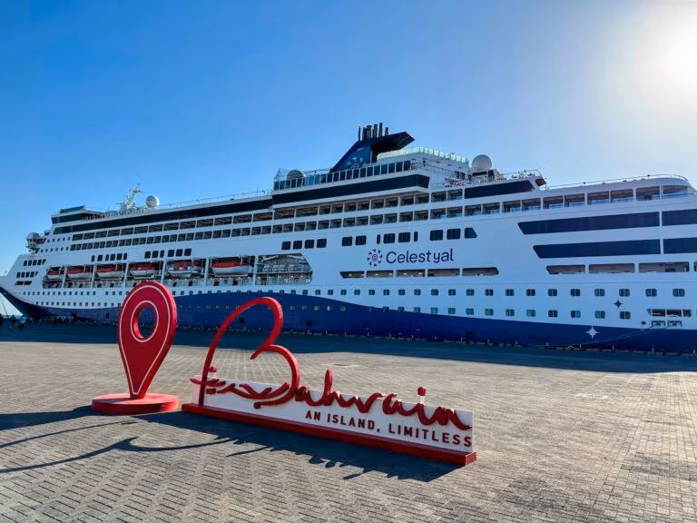 A large white and blue cruise ship, perfect for the Celestyal Journey Trip, is docked at a port. In the foreground, a red sign reads "Bahrain" with the slogan "An island, limitless" and features a location pin. The sky is clear and blue.
