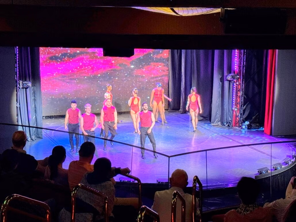 A group of performers in red costumes dances on a stage under a starry sky with pink clouds, capturing the essence of a Celestyal journey. The stage is set before an audience in a dimly lit theater, where the performers pose energetically, facing their captivated viewers.