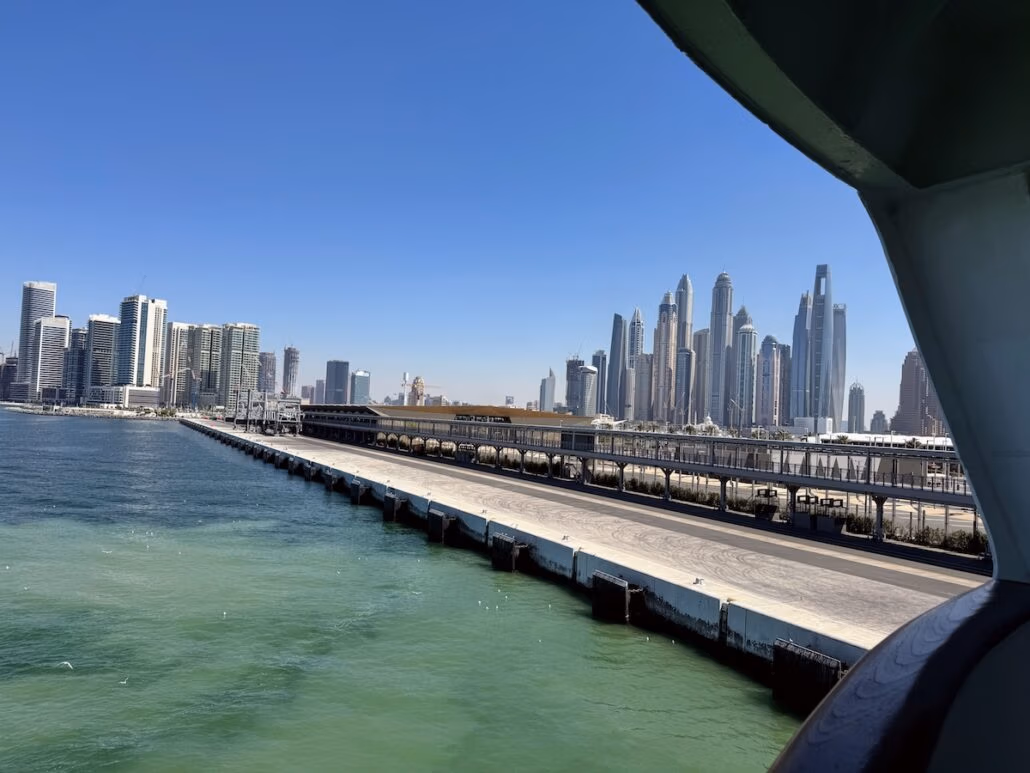 View of a harbor with calm green waters and a pier on the left side, reminiscent of embarking on a Celestyal Journey. In the distance, a cityscape with modern skyscrapers under a clear blue sky mirrors why this midsize ship feels perfect. Part of a structure is visible on the right side.