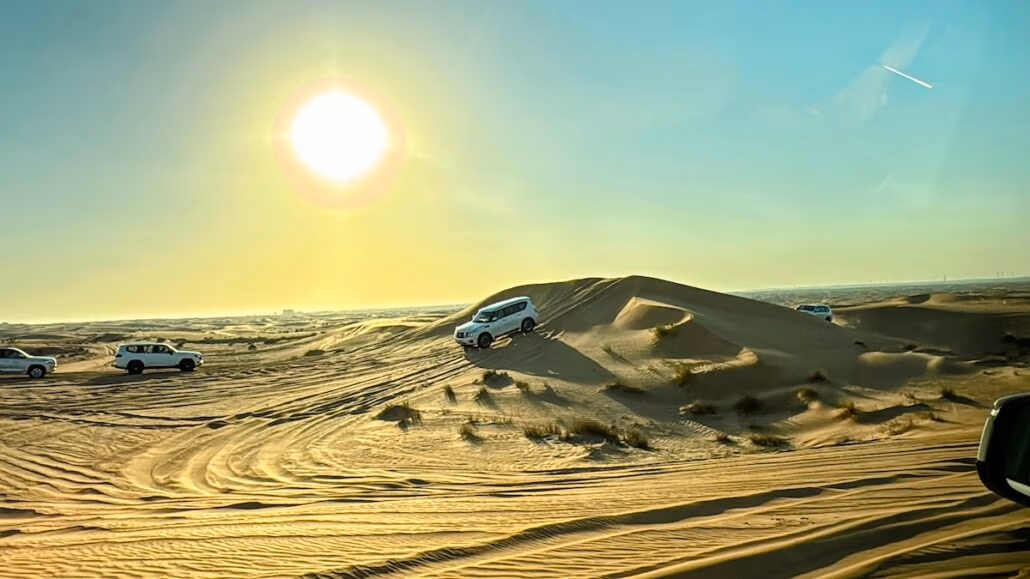Several SUVs traverse sandy dunes under a clear sky with the sun low on the horizon, reminiscent of a Celestyal journey. The smooth terrain bears wind patterns on the sand, echoing a perfect adventure.