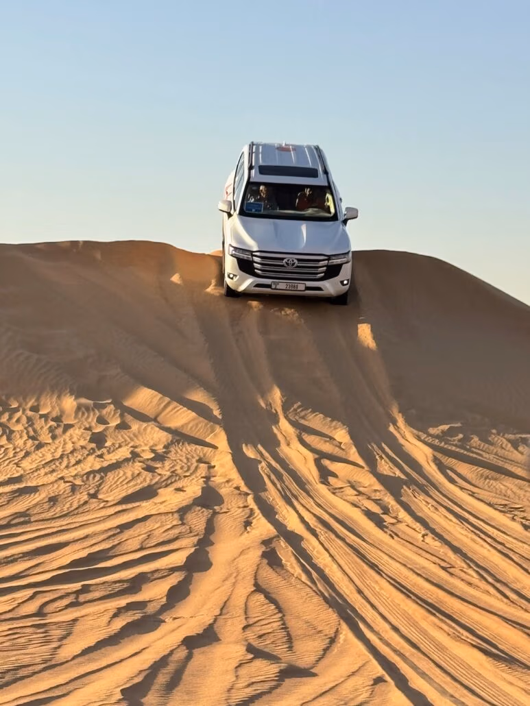 A white SUV is poised atop a sand dune under a clear blue sky, ready to descend. The sand, marked by the tracks of previous adventurers, exudes the thrill of off-road driving. Much like embarking on a Celestyal Journey, this scene captures the essence of exploration and discovery.