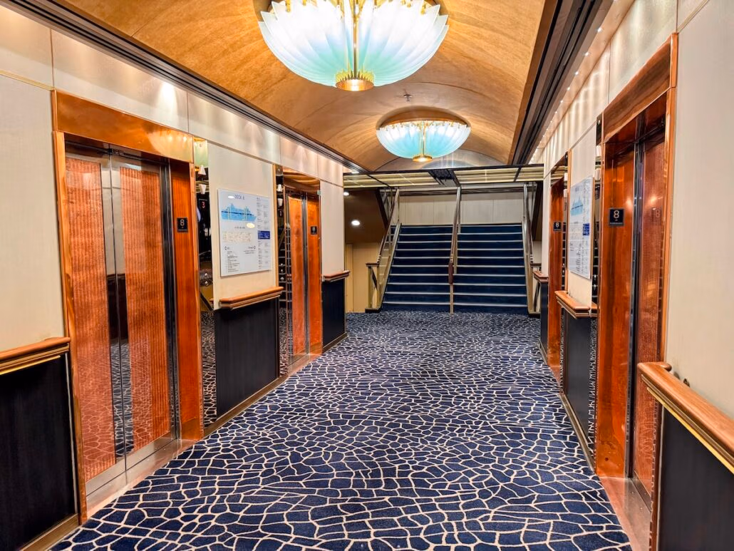 A corridor with three copper-framed elevators and a patterned carpet reminiscent of a Celestyal Journey. Two large, decorative ceiling lights illuminate the area. An emergency exit sign is visible near a staircase leading up at the end of the hallway, ensuring you're always prepared—a good thing on any adventure.