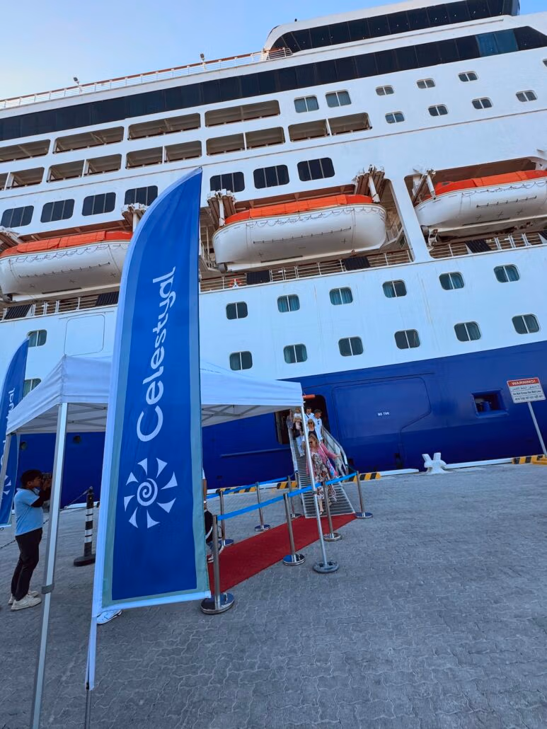 A large cruise ship, the Celestyal Journey, is docked at the port. Blue and white banners with "Celestyal" grace a red carpet leading up to the ship. Life rafts line its exterior under a clear sky—a typical cruise scene, if there ever was one.
