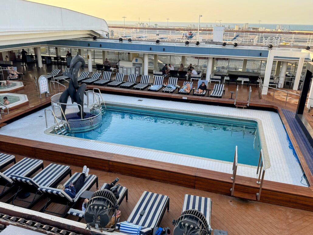 The pool area of the Celestyal Journey cruise features a rectangular swimming pool surrounded by striped lounge chairs, where guests relax. A dolphin statue graces the pool's corner, and the backdrop offers a stunning view of the ocean and clear sky.