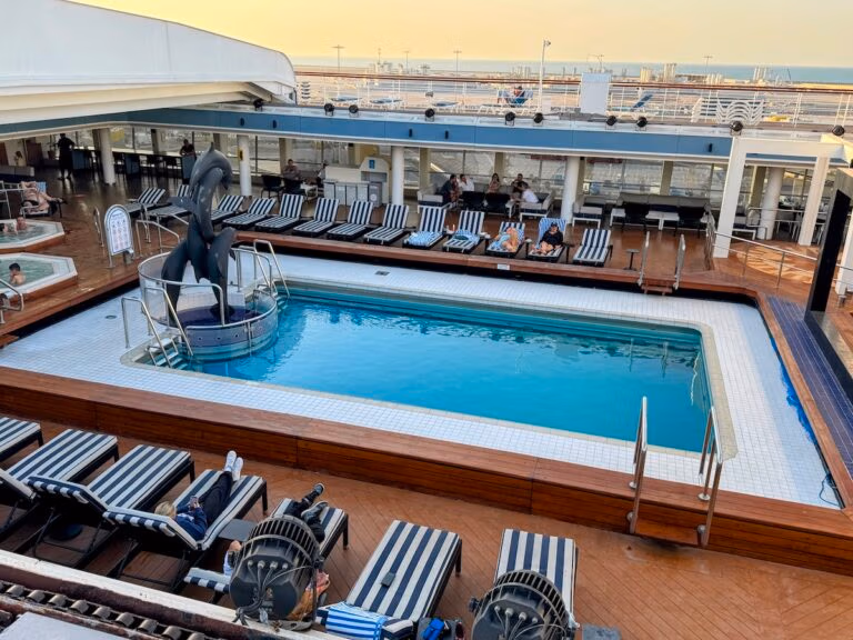 The pool area of the Celestyal Journey cruise features a rectangular swimming pool surrounded by striped lounge chairs, where guests relax. A dolphin statue graces the pool's corner, and the backdrop offers a stunning view of the ocean and clear sky.