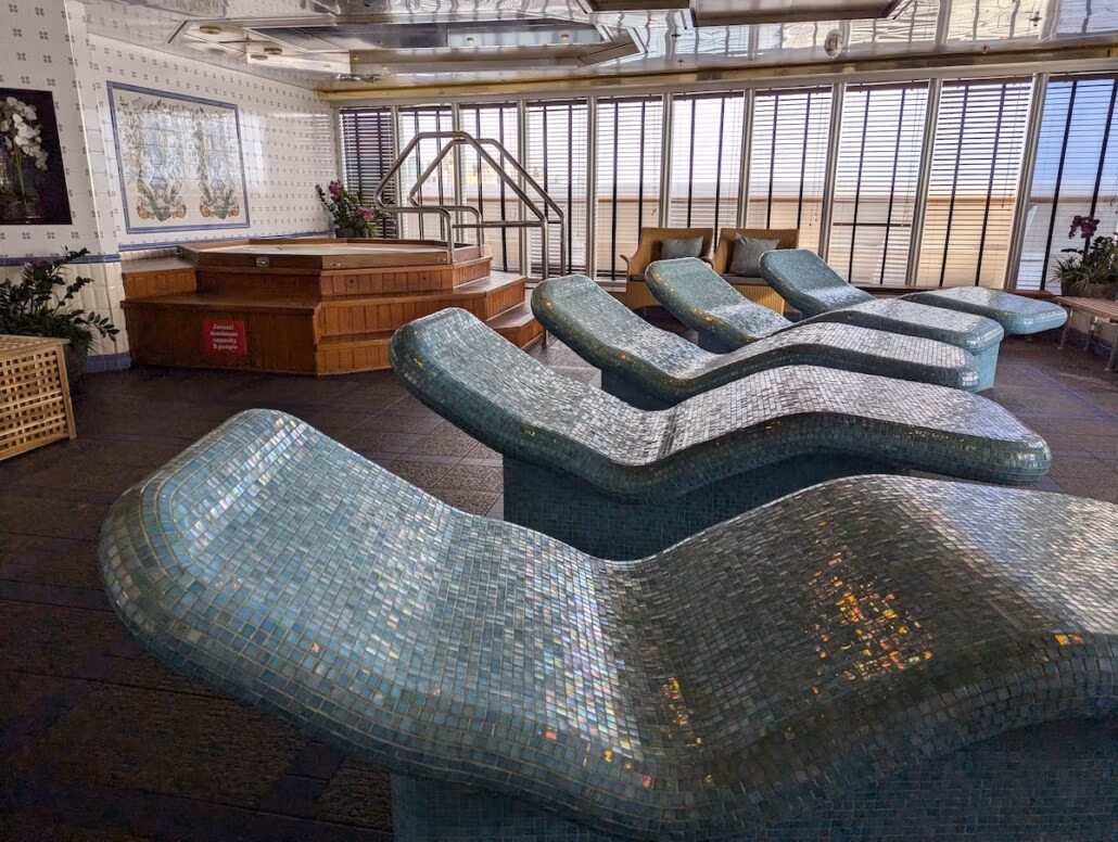 A spa room aboard the Celestyal Journey features several blue-tiled, curved lounge chairs and a wooden hot tub in the background. Wooden shutters line the windows, while a wicker basket and potted plants contribute to the serene decor—just one good thing about this typical cruise experience.