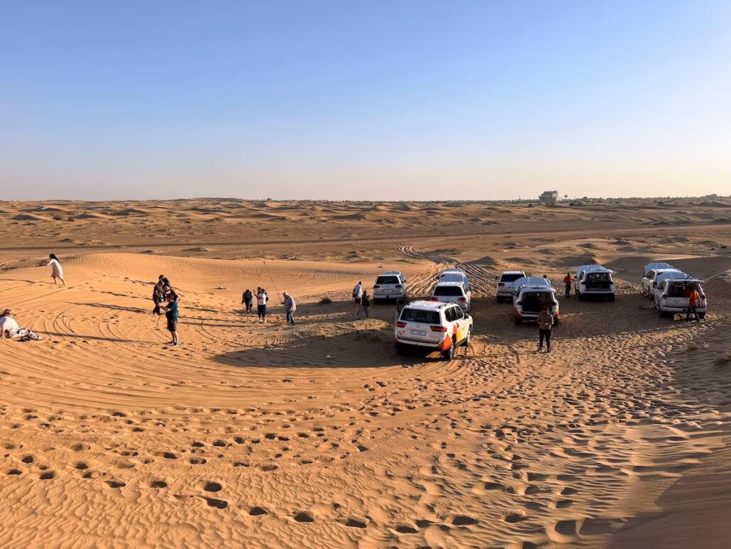 A group of people and several SUVs are parked on a sandy desert landscape under a clear sky, creating a unique experience. The vehicles and individuals are scattered on the dunes, with tire tracks visible in the sand as the horizon stretches into the distance.