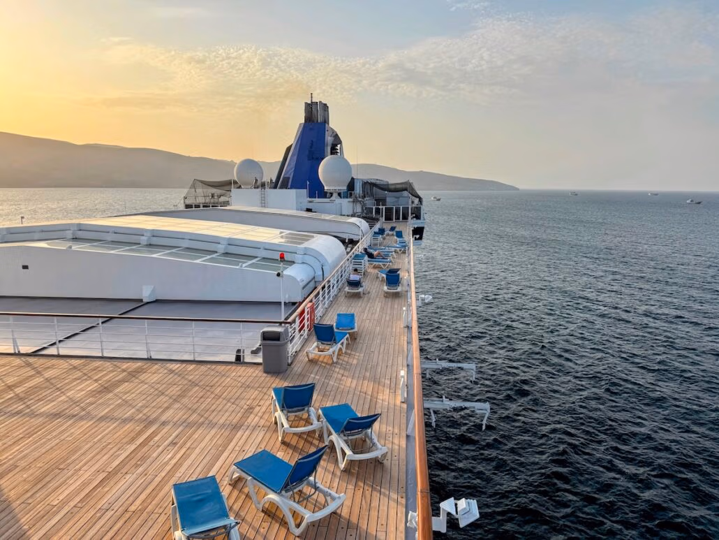 A wooden deck on the Celestyal Journey cruise ship features several blue lounge chairs and white side tables. The ship's structure, including radar domes and a blue funnel, is visible. Calm waters and a hazy horizon extend to the right under a partly cloudy sky—a typical cruise scene.