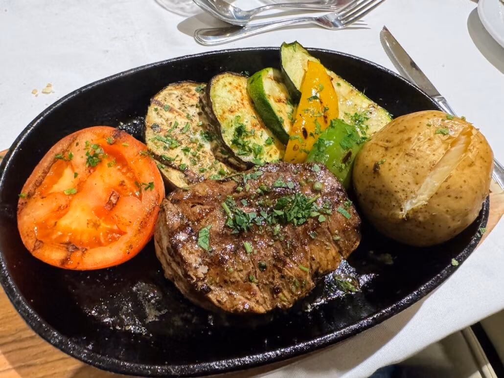 A sizzling steak served on a black platter alongside a roasted tomato slice, grilled zucchini, and a baked potato is truly a good thing. The dish is elegantly garnished with herbs, while a fork and knife rest on the white tablecloth in the background.