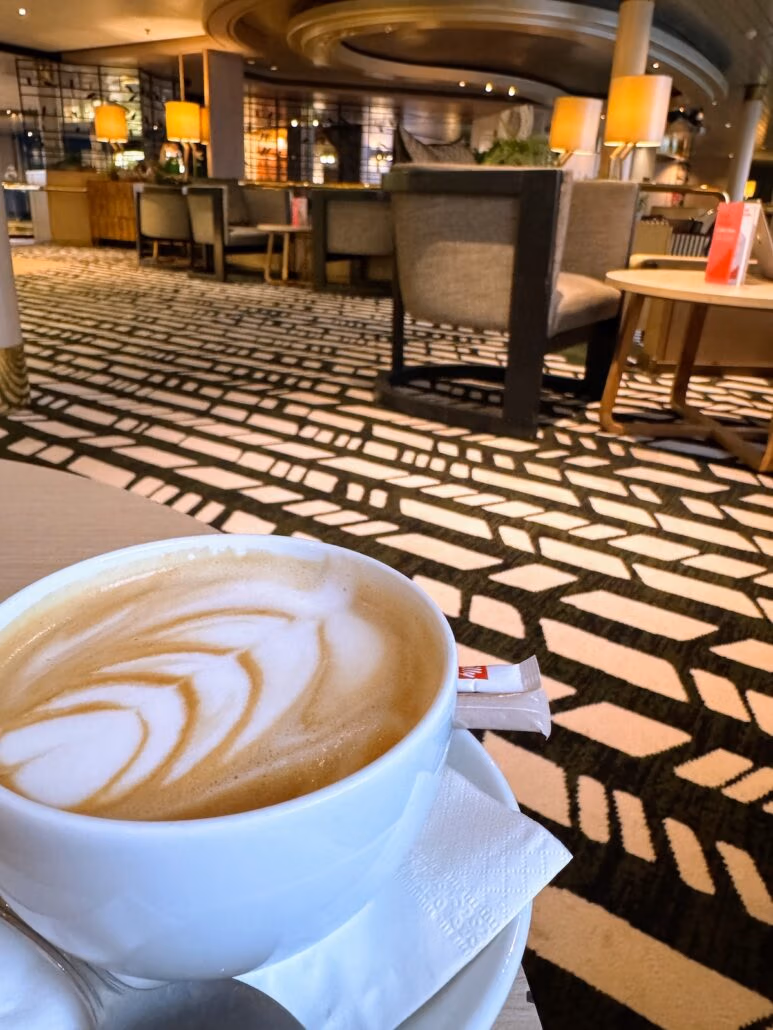 A close-up of a cappuccino with leaf-shaped latte art in a white cup on a napkin. The background, reminiscent of a Celestyal Journey, showcases a modern café with patterned carpet, wooden tables, gray chairs, and warm ambient lighting from lamps.