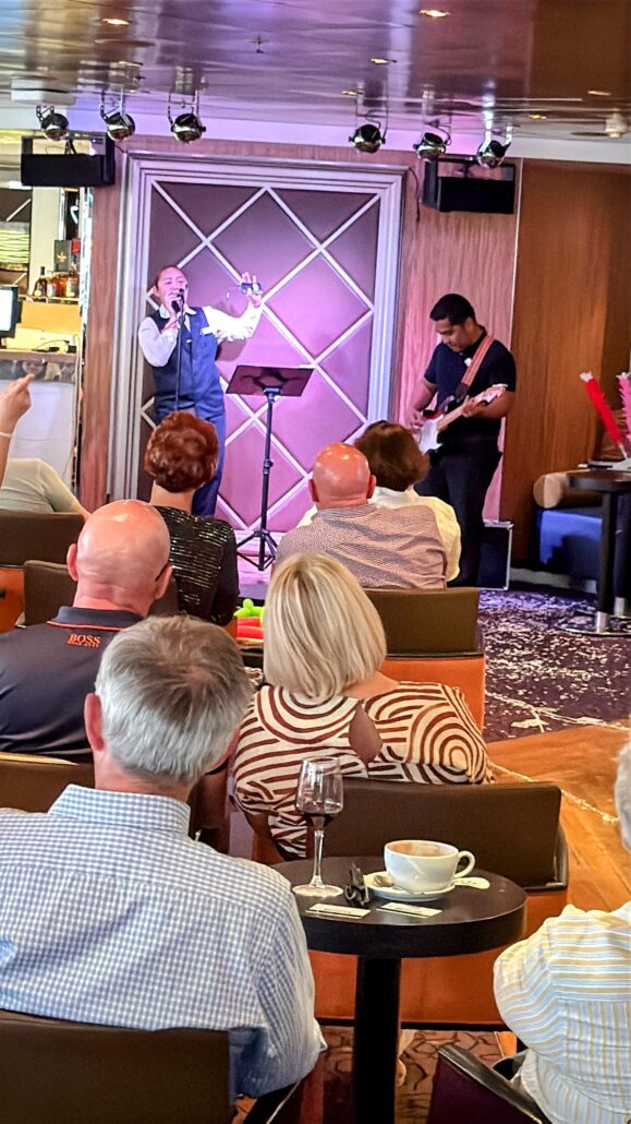 A singer and guitarist craft a unique experience on a small stage in a cozy venue. Audience members, seated with drinks at their tables, enjoy the warm lighting and decorative backdrop, akin to an intimate evening on a Celestyal Journey cruise.