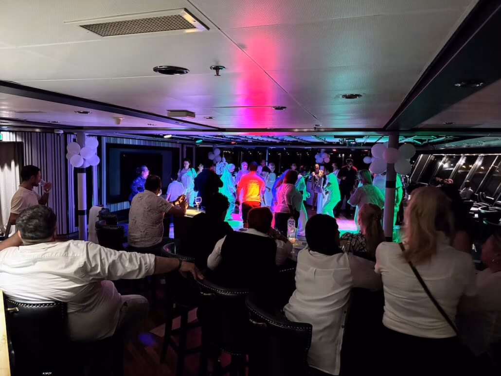 On a dimly lit dance floor, reminiscent of a Celestyal Journey, people are dancing under colorful lights. Others stand or sit around the perimeter, observing this captivating scene. White balloons adorn the walls, and recessed lighting on the ceiling adds to the charm—a voyage beyond a typical cruise experience.