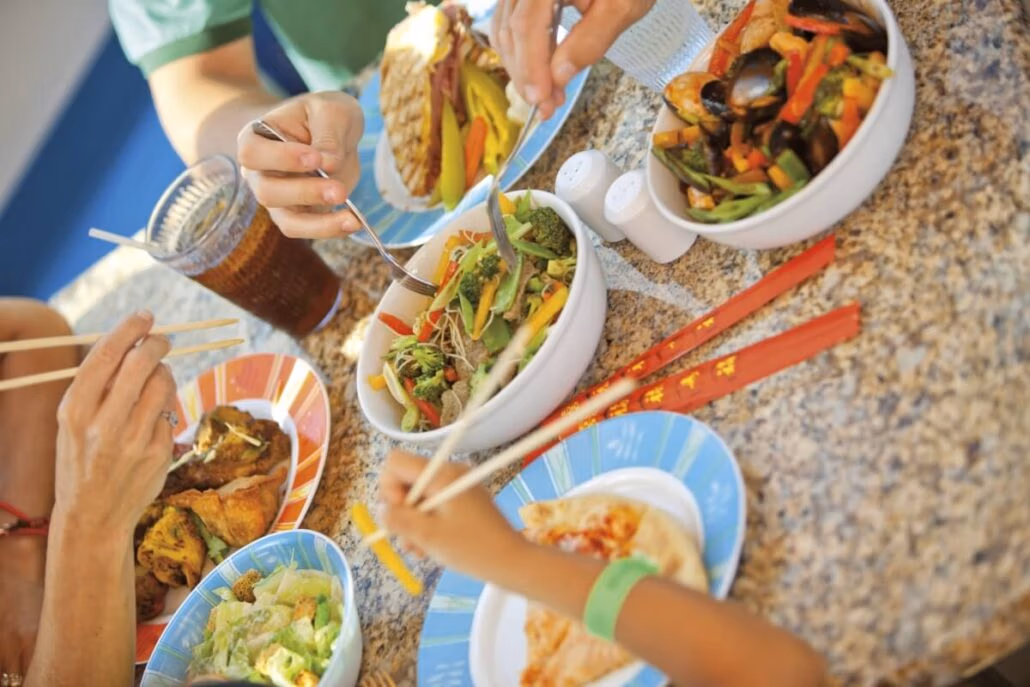 A group of people is enjoying a feast reminiscent of Carnival Cruise menus at a table adorned with stir-fried vegetables, salad, and meat. Plates, bowls, and chopsticks abound on the speckled surface, with a glass of dark liquid adding to the vibrant scene.