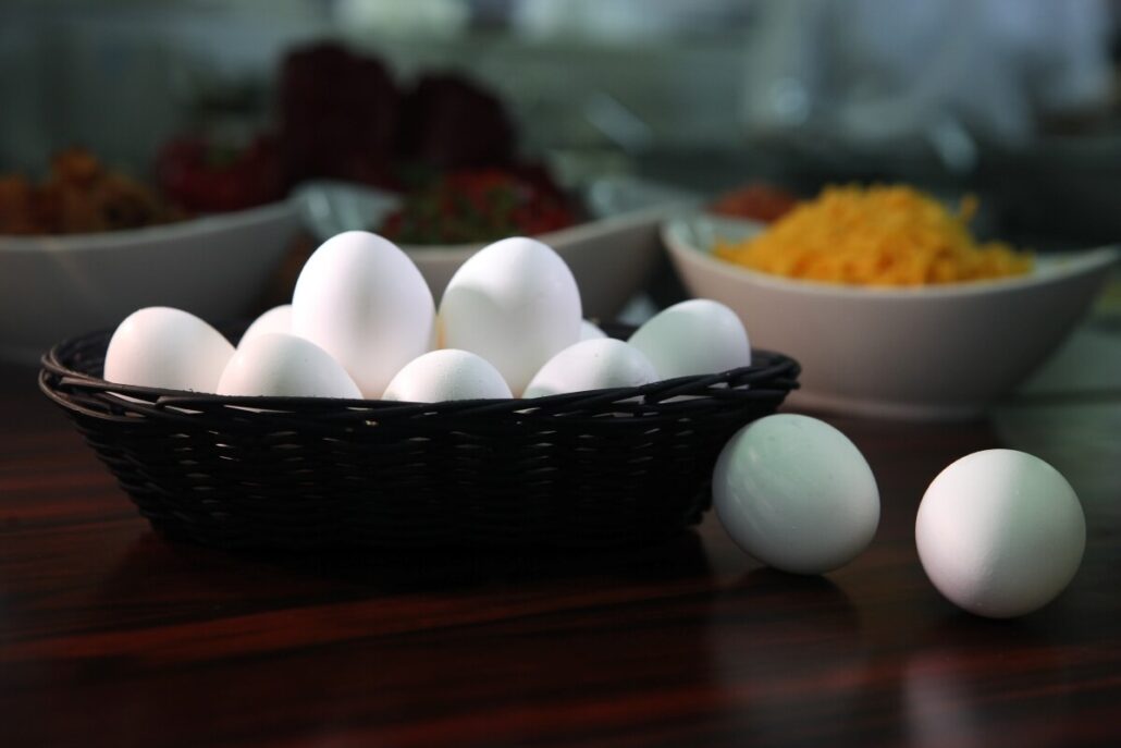 A wicker basket filled with white eggs sits atop a wooden table, two eggs resting outside it. Amidst whispers of an egg shortage, the blurred background reveals bowls brimming with ingredients like cheese and vegetables, reminiscent of a Carnival Cruise Line breakfast spread.