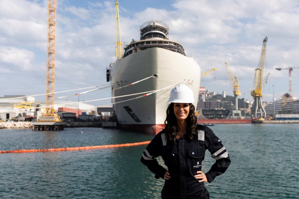 A person wearing a hard hat and safety gear stands in front of the Four Seasons Yacht docked at a bustling shipyard. Several cranes surround the area under a partly cloudy sky, their reflections shimmering alongside the yacht in the water.