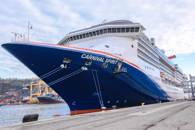 A large cruise ship named "Carnival Spirit" is docked at a port, its dark blue hull contrasting with the white superstructure. Nearby, cranes loom over another ship undergoing service under a partly cloudy sky.